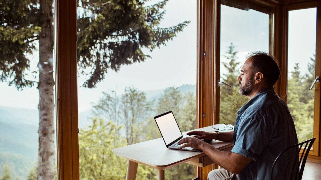 A person sitting at a desk typing on a computer looking out a window towards the water. 