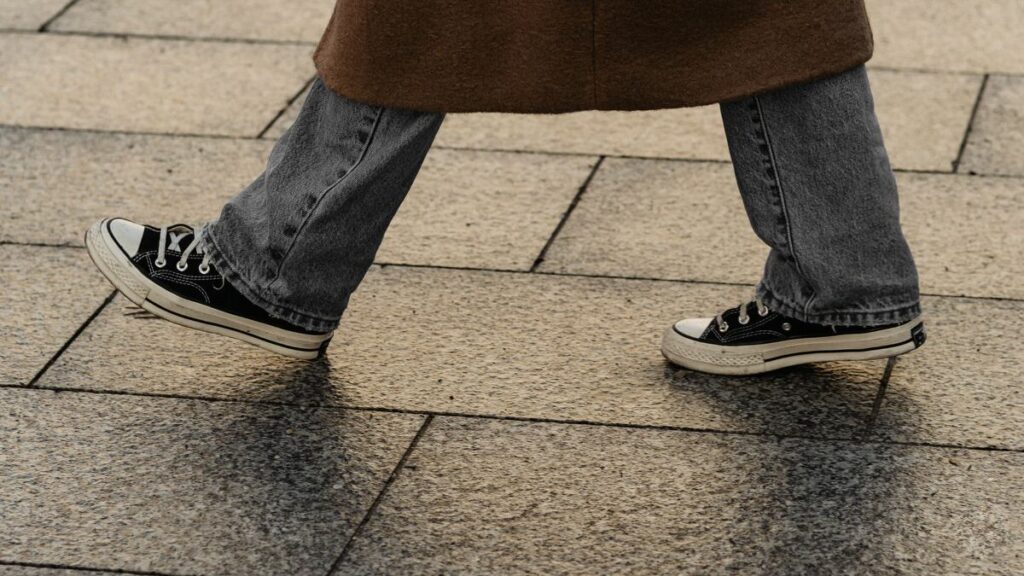 A close-up of someone's lower legs walking, featuring classic black and white Converse sneakers paired with grey denim jeans and a brown coat, on a textured sidewalk.