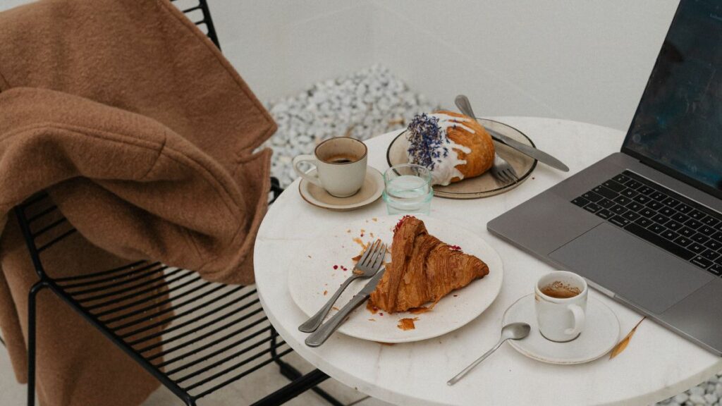 An overhead view of a cozy outdoor workspace with an unfinished croissant on a plate, a half-eaten glazed pastry, an empty espresso cup, and an open laptop on a white round table