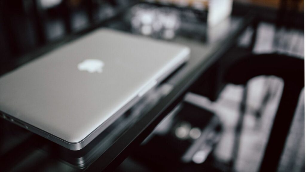 A closed MacBook on a reflective glass surface, with the iconic apple logo visible on the cover, set against a blurred background, emphasizing focus on the laptop