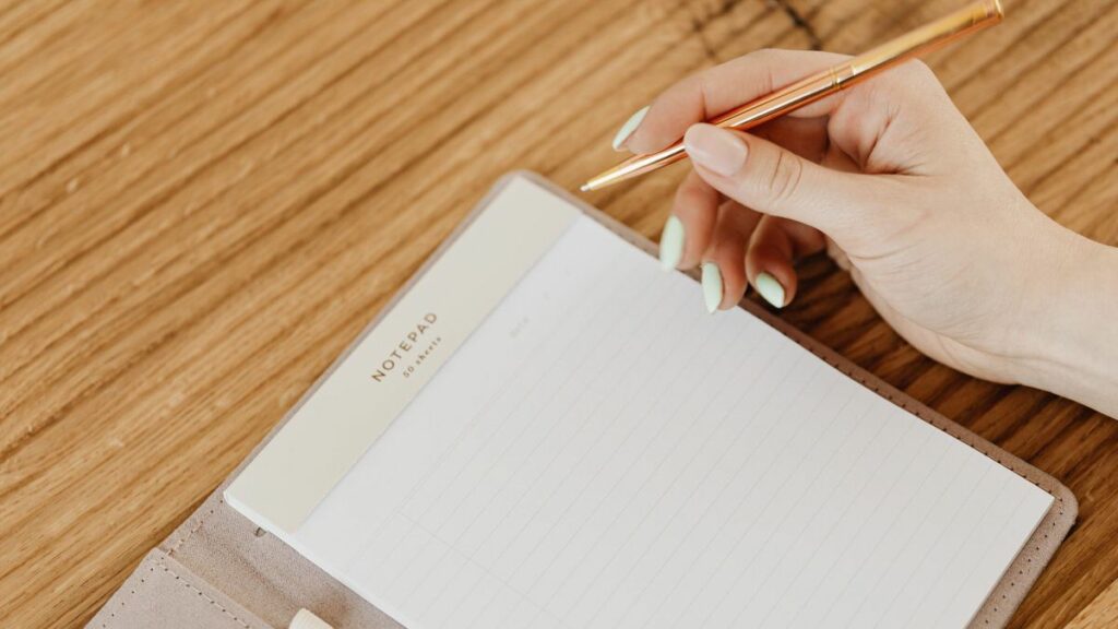 A person's hand holding a gold pen over a blank notepad on a wooden table, suggesting the beginning of writing or note-taking